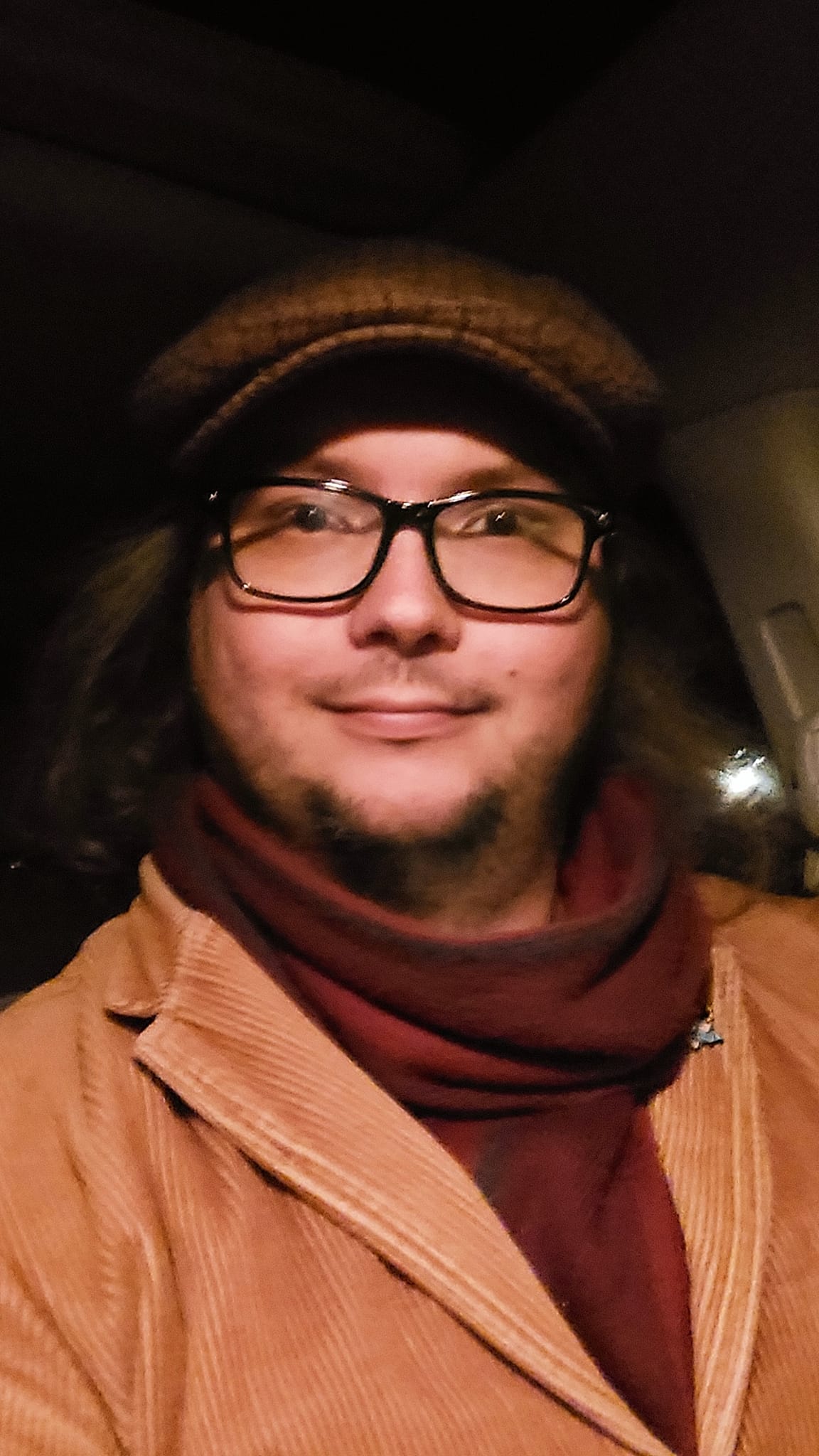 A man with long green hair wearing a brown corderoy coat and red scarf sits in a car after dark